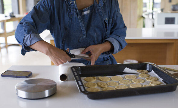 Mujer morena con delantal azul corta con tijeras papel manteca junto a ella hay una asadera o placa para horno con galletitas caseras, balanza y celular sobre la mesada de la cocina. 