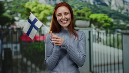 Woman smiling holding finland flag outdoors in city street showcasing finnish pride with redhead hair and joyful expression