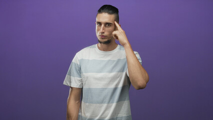 Fototapeta premium Man points finger to temple with right hand, standing in front of purple backdrop wearing striped tshirt in studio; thoughtful focus.