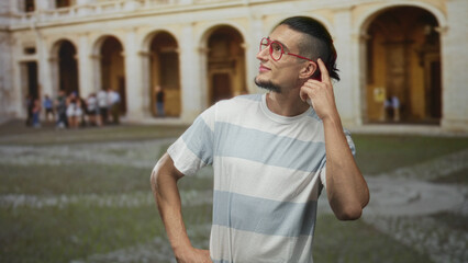 Man in striped white and blue t shirt with red glasses looking up, hand on hip in front of historic stone building courtyard; quiet curiosity.