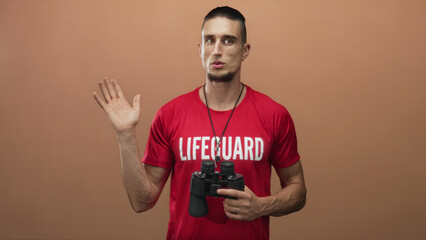 Man lifeguard waves raised left hand while holding binoculars and wearing red lifeguard shirt in studio; duty vigilance readiness.