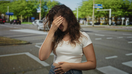 Woman in white crop top with hand on forehead on city street near crosswalk rubbing forehead for...