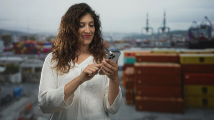 Woman texting on smartphone at port with stacked shipping containers and cranes in the harbor; digital connection contentment.