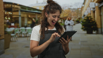 Woman in striped apron tapping tablet on restaurant terrace street while smiling and using both hands to type on screen; small business happiness.