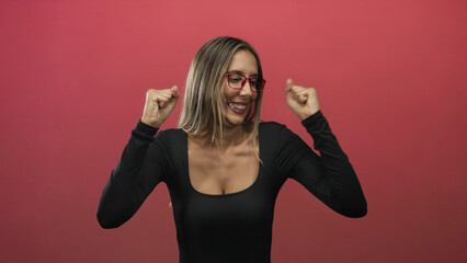Woman with fists raised in studio against a red pink backdrop, wearing red glasses, smiling while celebrating  joy celebration. © Krakenimages.com