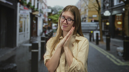 Fototapeta premium Blonde woman with clasped hands standing among urban storefronts on a wet cobblestone street; hope faith.