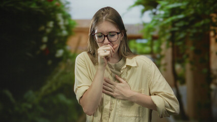 Young blonde woman wearing glasses with closed eyes clutches chest on sunlit forest path amid lush greenery; pain distress.