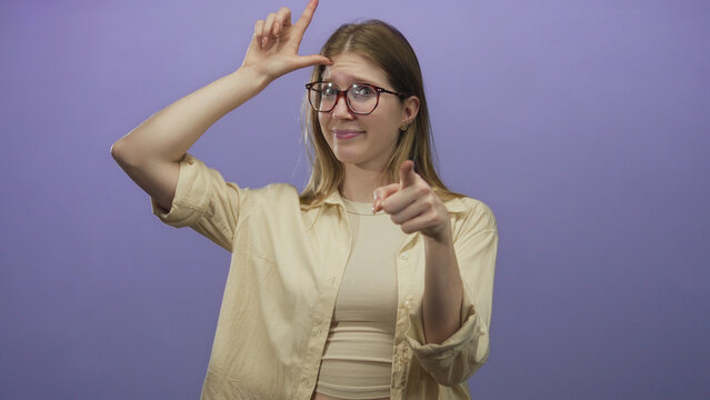 Blonde woman wearing glasses and beige top with finger forming l sign on forehead in purple studio; mockery.