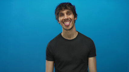 Young man sticking tongue out against a vivid blue studio wall showing the tongue gesture;...