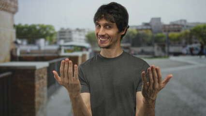 Young man in gray t shirt beckoning with outstretched bare hands on a busy city street beside brick wall; friendly invitation.