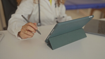 Middle age hispanic woman doctor holding stylus above tablet on white desk with laptop and stethoscope in clinic; calm care focus.
