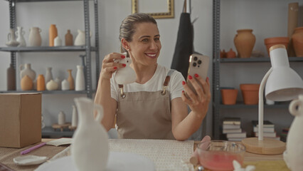 Middle age blonde woman holds ceramic vase to smartphone in pottery studio; craftsmanship passion joy.