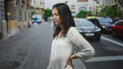 Fototapeta premium Woman standing with hand on lower back on a street beside cars and sidewalk, eyes closed and slight grimace of strain; backache discomfort.