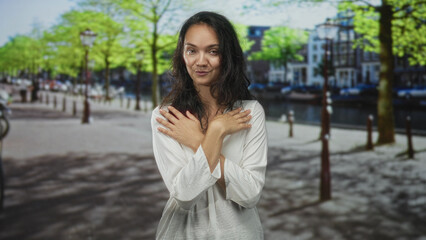 Fototapeta premium Woman with hand on cheek and crossed arms wearing a white blouse in a studio with canal street backdrop, soft smile and closed eyes; thoughtful.