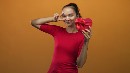 Woman holding red heart shaped gift box, showing peace sign fingers to eye, smiling in an orange studio backdrop; playful affection.
