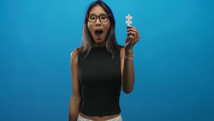 Thai woman wearing glasses and black top holding white puzzle piece with open mouth in studio;...