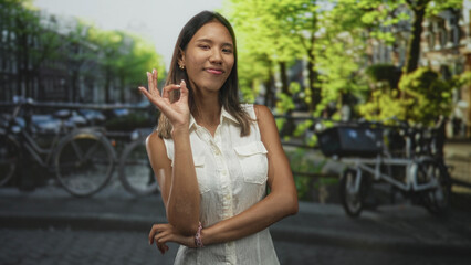 Fototapeta premium Thai woman makes ok hand gesture with visible bare hand and crossed arms on a street lined with bicycles and canal buildings; approval.