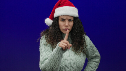 Hispanic woman in santa hat and sweater pointing finger in warning against a blue background, conveying disapproval and holiday authority.