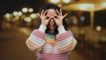 Fototapeta premium Woman making glasses gesture with hands, smiling joyfully in colorful sweater on an illuminated street at night, conveying playful outdoor vibes.