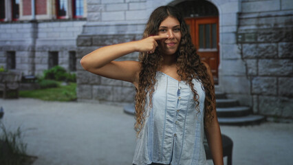 Obraz premium Hispanic teenage woman points finger to nose while smiling, sleeveless blue shirt visible, long curly hair, standing by stone building steps and wooden door; playful confidence.
