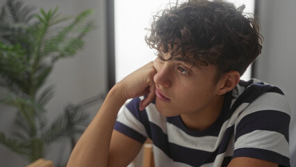 Teen boy with curly brown hair resting chin on hand, wearing striped shirt and leaning at table in studio; quiet pensive reflection.