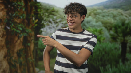 Fototapeta premium Man points finger to the right in a forest setting, smiling while wearing round glasses and a navy and white striped shirt by a tree trunk; joy.