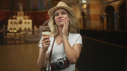 Woman holding ice cream cone, hand on cheek, wearing straw hat and camera around neck in building; toothache annoyance travel.