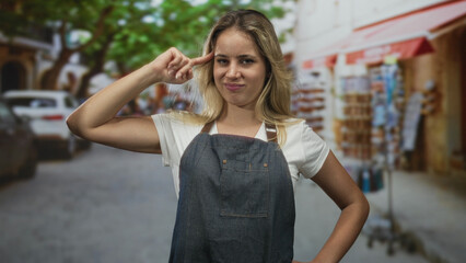 Fototapeta premium Woman points finger to temple on a busy street market wearing denim apron and white tshirt with one hand on hip and smirk visible; defiance.