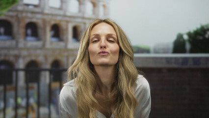 Portrait of a confident woman lips blowing salute before ancient coliseum roman blonde explorer posing amidst busy outdoors street heart shape moment.