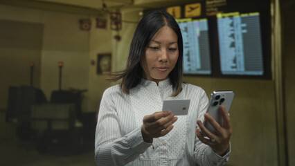 Woman holds creditcard and smartphone at busy airport terminal while checking flight details; calm travel planning.