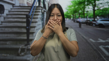 Fototapeta premium Woman with hands covering mouth on a street by stone steps and parked cars, wearing apron and bracelet; silence.