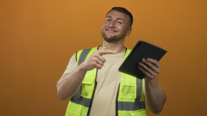 Man engineer in high visibility vest pointing finger at tablet and holding device in orange studio, smiling at camera; confidence productivity.