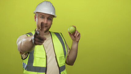 Man engineer in white hardhat and high visibility vest pointing finger while holding green apple in...