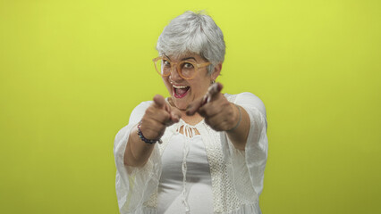 Woman with grey hair and glasses points both fingers at camera in studio while smiling and laughing, wearing white blouse; playful confidence.