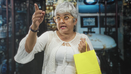 Woman, senior with grey hair and orange glasses, plus size, holding bright yellow shopping bag and points finger upward inside museum building while looking alert; curiosity discovery.