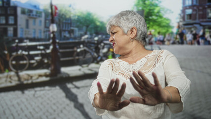 Woman with grey hair pushes palms forward in a stopping gesture toward bicycles on a cobblestone street; rejection caution.