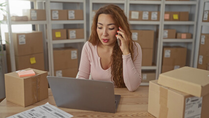 Woman holds phone to ear while using laptop and organizing parcels on desk in a building; small...