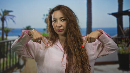 Woman points both index fingers down at her pink shirt on a building balcony with ocean view; confidence assertiveness.