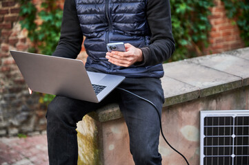 Cropped view of man sits on stone bench in front of brick wall, working on laptop and smartphone, connected to solar panel. Urban setting, casual attire highlight modern sustainable, mobile workspace.
