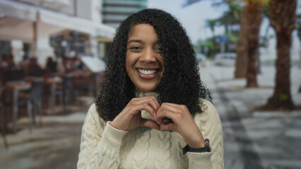 Smiling black woman makes heart shape with hands on sunny outdoor restaurant terrace with wide...