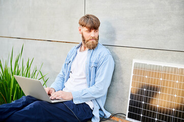 Bearded man works on laptop on lap. Solar panel nearby, emphasizing sustainable, modern eco-friendly workspace that combines technology and sustainability.