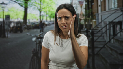 Woman with clenched fist near mouth and repeated hand gestures to ear, visible expressive face while standing on a street; frustration communication listening.