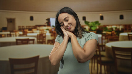 Woman clasping hands to cheek in sleeping gesture amid round tables inside banquet building; gentle rest serenity.