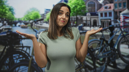 Woman shrugs with palms up in front of a bicycle and canal backdrop in a studio, raised hands and slight head tilt show indecision; uncertainty.