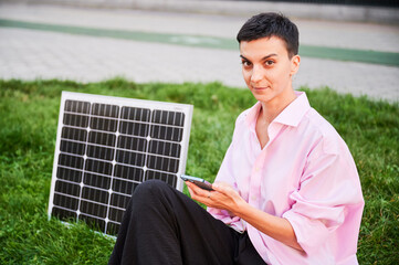 Happy woman using smartphone connected to photovoltaic solar panel. Integration of sustainable renewable energy into everyday life, demonstrating practical use of solar power for charging devices.