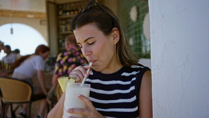 Young brunette woman sipping a creamy pineapple cocktail while holding a straw at a bar table with...