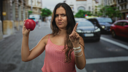 Young hispanic woman points finger to red apple while holding it near chest on a busy city street with cars behind; assertive caution.