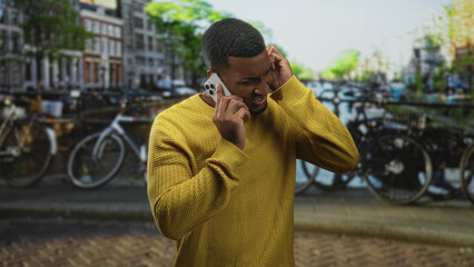 Young black man holds smartphone to ear and cups other ear with hand on street near canal and bicycles; frustration urgency.
