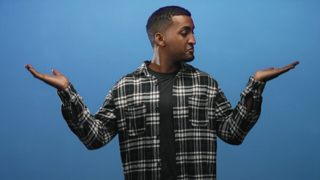 Young man wearing plaid shirt holds both open palms up in a questioning shrug pose against a blue backdrop in studio; uncertainty.