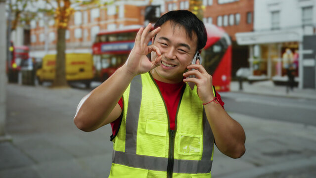 Man in safety vest making phone call with smartphone while smiling and gesturing okay sign on busy urban street with iconic red double-decker bus and delivery vans visible.
